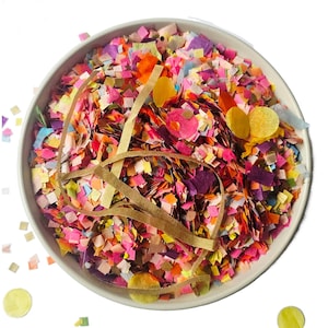 Peut inclure: Un bol blanc rempli de confettis colorés de différentes formes et tailles. Les confettis comprennent des carrés, des cercles et des cœurs dans des tons de rose, orange, jaune, violet et vert. Des rubans en papier brun sont également présents.