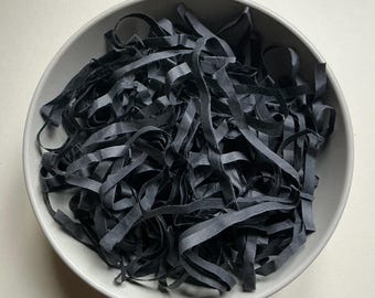 May include: A light gray ceramic bowl filled with a dense pile of thin, matte black paper strips. The strips are folded and tangled, creating a textured, layered appearance within the circular container. The bowl has a smooth, solid rim.