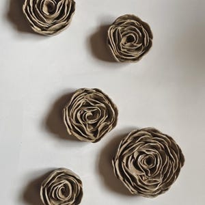 May include: Five individual ceramic flower sculptures shaped like roses in a neutral taupe colour. Each piece features layered, ruffled petals with a glossy finish, arranged in varying sizes from small to large, showcasing intricate, hand-formed details.