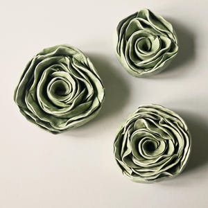 Puede incluir: Tres esculturas de rosas de cerámica verde salvia. Cada escultura presenta pétalos en capas que forman un centro en espiral. Las piezas están dispuestas sobre un fondo neutro, mostrando la textura y forma detalladas.