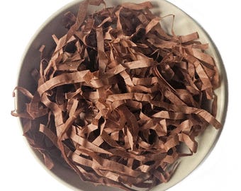 May include: A top-down view of a beige bowl filled with shredded brown paper. The paper is cut into long, thin strips, creating a textured, layered appearance. The bowl is round with a slightly raised rim, and the background is white.
