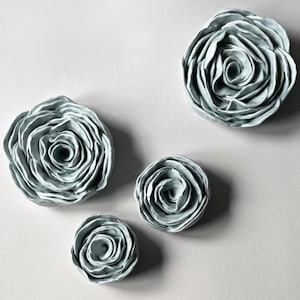 May include: A set of four pale blue ceramic flower sculptures arranged in a cluster. Each piece features layered, ruffled petals that spiral towards a central point, creating a dimensional, blooming rose-like appearance with a smooth finish.