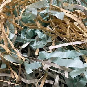 Op de afbeelding: Close-up van versnipperd papier in tinten beige, lichtgroen en grijs. Het papier is in lange, dunne stroken gesneden, waardoor een getextureerd, gelaagd effect ontstaat. Ideaal voor verpakking en cadeauverpakking.