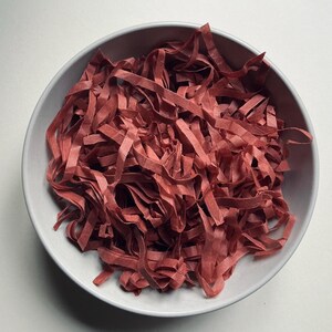 May include: A round, light grey bowl filled with a generous amount of thin, crinkled strips of dark red paper. The paper strips are tangled together, creating a textured, layered appearance within the smooth, circular container.