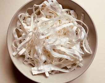 May include: A round, light brown ceramic bowl filled with a pile of thin, white, flat, ribbon-like paper strips. The strips are arranged in a loose, tangled heap, filling the interior of the bowl with a textured, layered appearance.