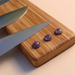 Puede incluir: Primer plano de una tabla de cortar de bamb&uacute; con tres imanes de piedras preciosas moradas y varios cuchillos de acero inoxidable. Los imanes tienen un borde de metal plateado. Los cuchillos son afilados.
