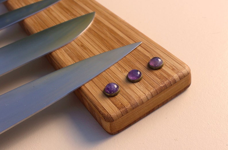 Puede incluir: Primer plano de una tabla de cortar de bamb&uacute; con tres imanes de piedras preciosas moradas y varios cuchillos de acero inoxidable. Los imanes tienen un borde de metal plateado. Los cuchillos son afilados.