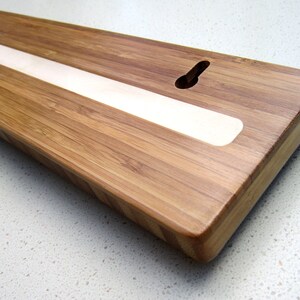 Puede incluir: Una tabla rectangular de madera de bamb&uacute; con una tira clara empotrada en el centro. La tabla tiene un recorte en forma de ojo de cerradura para colgar. Los bordes son redondeados.