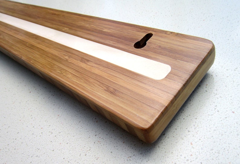 Puede incluir: Una tabla rectangular de madera de bamb&uacute; con una tira clara empotrada en el centro. La tabla tiene un recorte en forma de ojo de cerradura para colgar. Los bordes son redondeados.