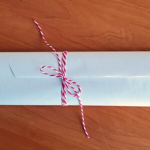 Puede incluir: Un paquete rectangular envuelto en papel claro, atado con hilo rojo y blanco. El paquete est&aacute; sobre una superficie de madera. El hilo est&aacute; atado en un lazo.