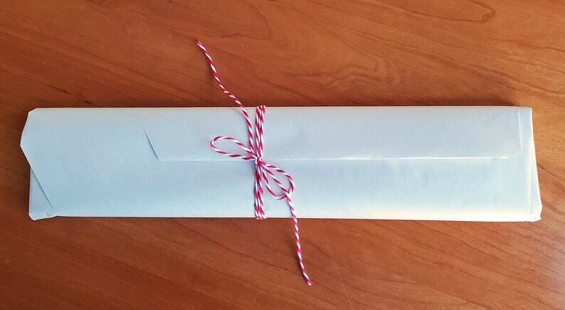 Puede incluir: Un paquete rectangular envuelto en papel claro, atado con hilo rojo y blanco. El paquete est&aacute; sobre una superficie de madera. El hilo est&aacute; atado en un lazo.