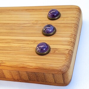 Puede incluir: Tres cabujones morados con efecto veteado, engastados en biseles de lat&oacute;n antiguo. Las piedras est&aacute;n dispuestas sobre una tabla de cortar de bamb&uacute;, mostrando su color y dise&ntilde;o &uacute;nicos. Los cabujones son una hermosa adici&oacute;n a cualquier colecci&oacute;n de joyas.