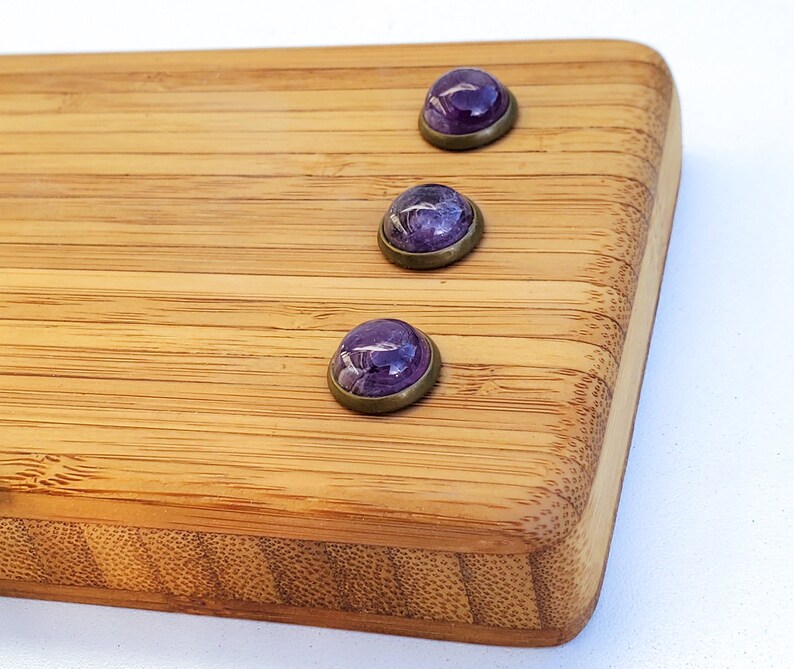 Puede incluir: Tres cabujones morados con efecto veteado, engastados en biseles de lat&oacute;n antiguo. Las piedras est&aacute;n dispuestas sobre una tabla de cortar de bamb&uacute;, mostrando su color y dise&ntilde;o &uacute;nicos. Los cabujones son una hermosa adici&oacute;n a cualquier colecci&oacute;n de joyas.