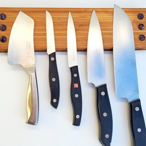 Puede incluir: Un soporte de cuchillos de bamb&uacute; con cinco cuchillos de acero inoxidable. Los cuchillos tienen mangos negros y est&aacute;n dispuestos horizontalmente. El soporte tiene tres acentos decorativos morados.