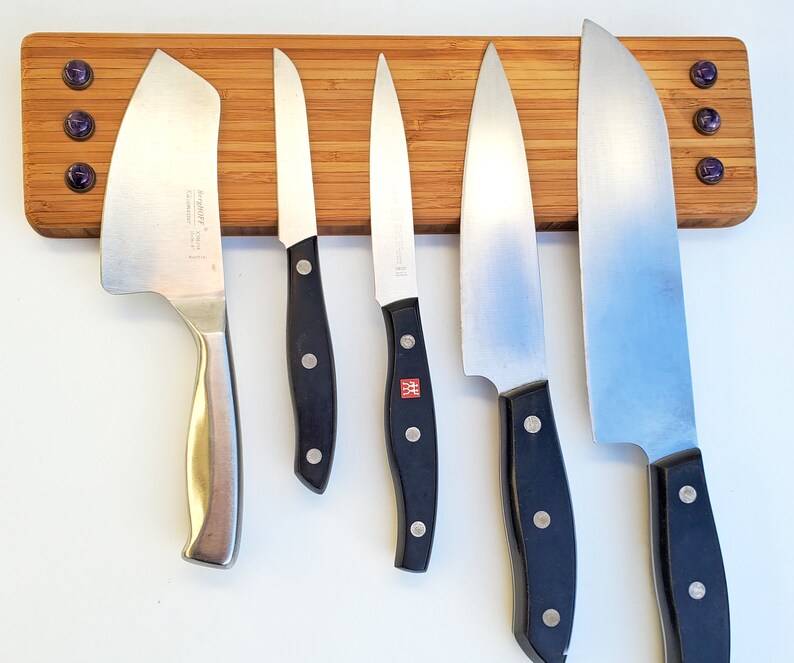 Puede incluir: Un soporte de cuchillos de bamb&uacute; con cinco cuchillos de acero inoxidable. Los cuchillos tienen mangos negros y est&aacute;n dispuestos horizontalmente. El soporte tiene tres acentos decorativos morados.