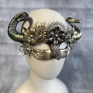May include: A decorative mask with a horn-like design. The mask features a combination of gold, silver, and green tones. Intricate floral and leaf embellishments adorn the mask's surface, creating a detailed and ornate appearance. The mask is displayed on a mannequin head.