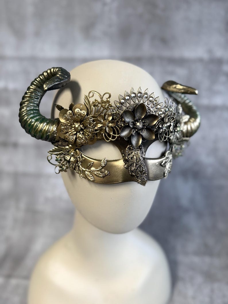 May include: A decorative mask with a horn-like design. The mask features a combination of gold, silver, and green tones. Intricate floral and leaf embellishments adorn the mask's surface, creating a detailed and ornate appearance. The mask is displayed on a mannequin head.