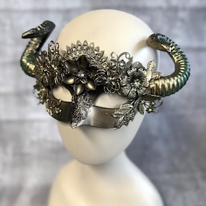 May include: A decorative mask with a metallic finish, featuring ornate floral embellishments and curved horns. The mask has a central eye covering and is adorned with intricate details, including rhinestones and leaf accents. The horns are painted with a green and gold gradient.