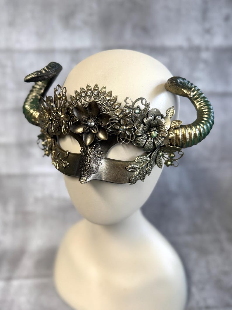 May include: A decorative mask with a metallic finish, featuring ornate floral embellishments and curved horns. The mask has a central eye covering and is adorned with intricate details, including rhinestones and leaf accents. The horns are painted with a green and gold gradient.