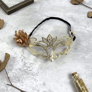 May include: A gold-colored masquerade mask with a floral accent and a black elastic band. The mask features a decorative filigree design with clear rhinestones. A small glass bottle filled with gold glitter is also present.