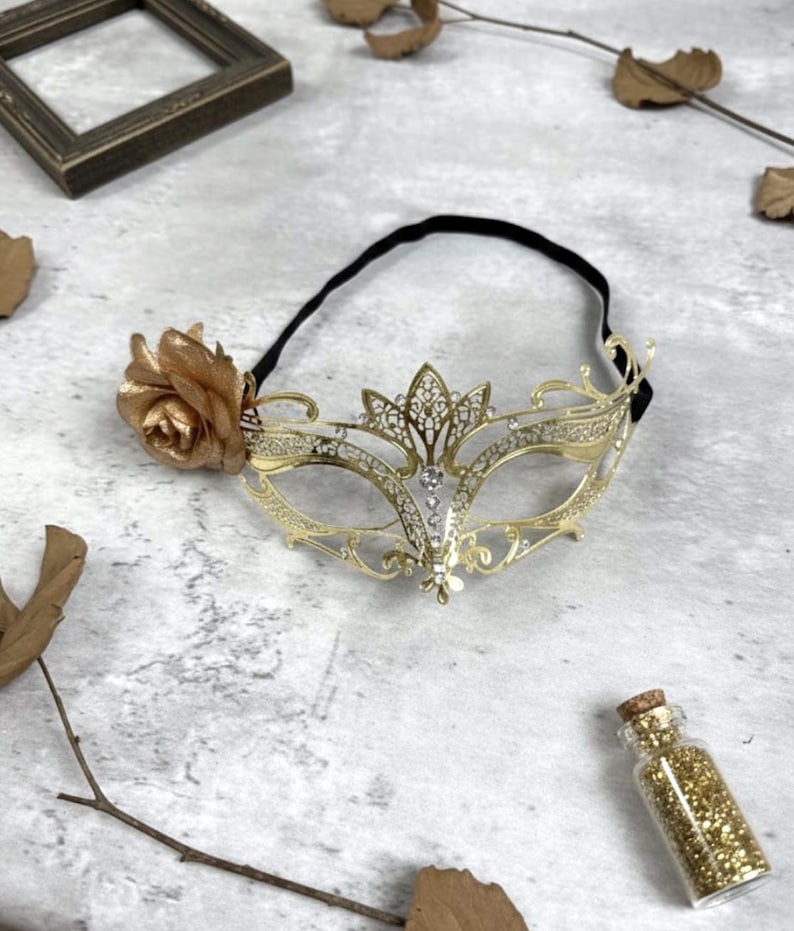 May include: A gold-colored masquerade mask with a floral accent and a black elastic band. The mask features a decorative filigree design with clear rhinestones. A small glass bottle filled with gold glitter is also present.