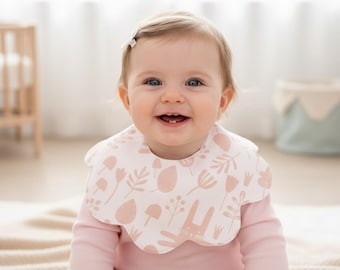 Può includere: Un bambino indossa un body rosa a maniche lunghe e un bavaglino bianco con bordo smerlato. Il bavaglino presenta un motivo ripetuto di fiori rosa chiaro, foglie e un coniglietto. Il bambino sorride.