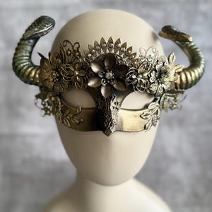 May include: A decorative mask with a gold finish, featuring curved horns and floral embellishments. The mask has a central eye covering and is adorned with intricate metal flowers and leaf details. The horns have a green and gold gradient.