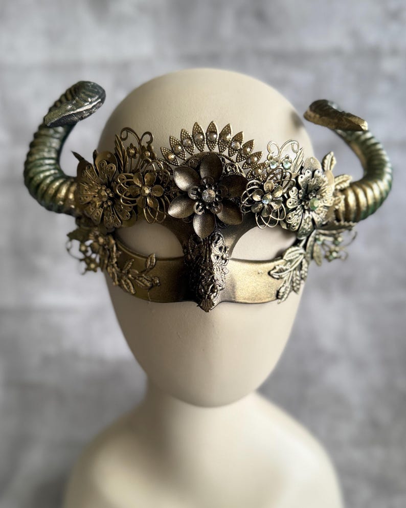 May include: A decorative mask with a gold finish, featuring curved horns and floral embellishments. The mask has a central eye covering and is adorned with intricate metal flowers and leaf details. The horns have a green and gold gradient.