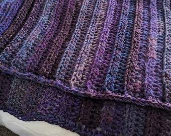 May include: A hand-crocheted blanket featuring a ribbed texture and a variegated color palette of deep purple, violet, and dark blue tones. The yarn displays subtle flecks of lighter shades throughout the dense, repeating stitch pattern.