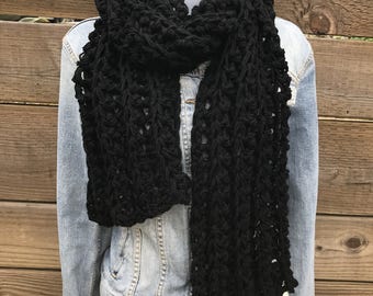May include: A thick, black, chunky knit scarf with a prominent vertical ribbed texture, draped around the neck of a mannequin wearing a light blue denim jacket over a cream-colored skirt. The scarf features a loose, open-stitch construction.