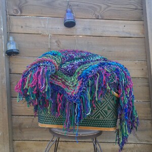 May include: A colorful crocheted throw blanket with long fringe hangs over a wicker basket. The blanket is a mix of purple, blue, green, and pink yarn.