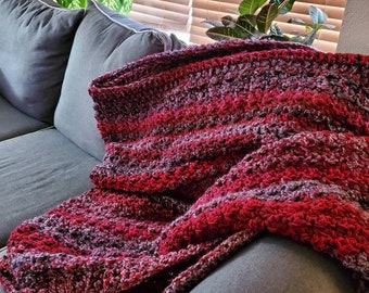 May include: A handmade crochet blanket in shades of red, burgundy, and purple, draped over a gray sofa. The blanket's texture is visible, with a mix of light and dark hues. The cozy throw blanket is perfect for home decor.