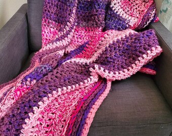 May include: A crocheted throw blanket in shades of pink and purple. The blanket is made with a chunky yarn and has a textured stitch pattern.