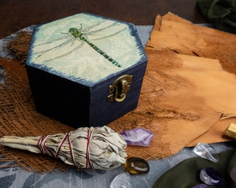 Puede incluir: Una caja de madera hexagonal con un diseño de libélula en la tapa. La caja está pintada de azul y tiene un cierre de latón. La caja está rodeada de salvia, cristales y cuero.