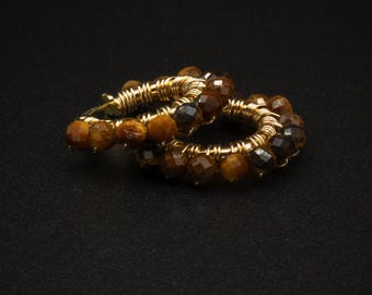 May include: Two gold-colored hoop earrings with faceted, brown tiger eye beads. The beads are strung around the hoops, which are wrapped with gold wire. The earrings are displayed on a black background.