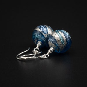 May include: A pair of handcrafted earrings with blue glass beads and silver leaf detailing. The beads are suspended from sterling silver ear wires. The earrings are set against a black background, highlighting the jewellery's details.
