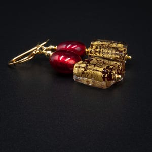 May include: A pair of drop earrings featuring a gold-toned hook, a smooth deep red oval bead, and a square glass bead infused with metallic gold leaf flakes. The design incorporates gold-colored metal accents connecting the beads in a linear arrangement.