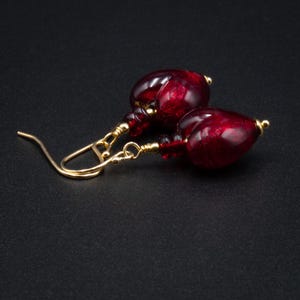 May include: A pair of drop earrings featuring deep red, translucent glass beads with a textured, marbled appearance. The teardrop-shaped beads are attached to gold-toned metal ear wires with delicate wire wrapping and small gold-toned metal accents.