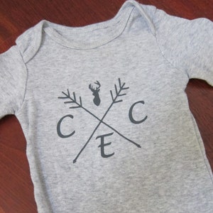May include: A gray baby onesie with a black deer silhouette and crossed arrows with the letters "C E C" printed on the front.