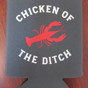 May include: Gray neoprene can cooler with white text that reads "Chicken of the Ditch" and a red silhouette of a crawfish.