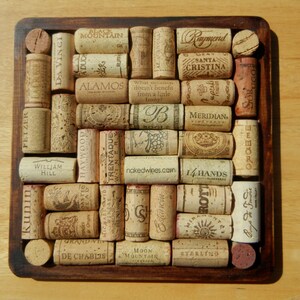 May include: A square wooden coaster with a grid of wine corks. The corks are arranged in a pattern and have various labels, including "Raymond", "Santa Cristina", "Meridian", "14 Hands", "Moon Mountain", and "Sterling".