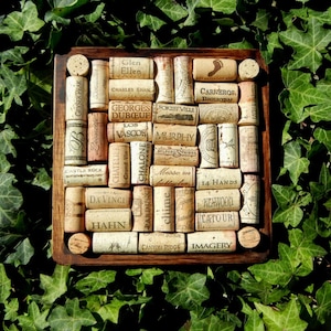 May include: A square wooden coaster with a grid of wine corks, each with a different winery name printed on it. The names include: Geyser Peak, Leonetti, Chateau, Grand Vin, Chalone, Parducci, Davinci, Hahn, Chime, Canyon Ridge, Castle Rock, OB, Sollicille, Charles Shaw, Georges Duboeuf, Los Vascos, Muirphy, Belford Spriegs, Messo in, 14 Hands, Latour, Imagery, Duhart Milou, Carneros, Bighorn, and Forestville.