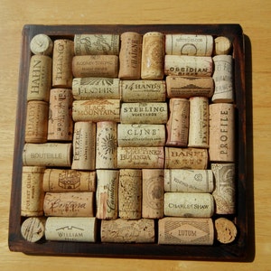 May include: A square wooden coaster with a grid of wine corks. The corks are arranged in a pattern and have various labels, including "FOREST GLEN", "TIZIANO", "QUADY", "HAHN", "MADERA", "CAM", "BOULEA", "FETZER", "WIS", "BOUTEILLE", "Bonfassa", "WILLIAM", "Cambria", "RODNEY STRONG", "CHAIGNE", "odirWELCOM", "LOBSIDIAN", "JLOHR", "14 HAND'S", "ERBE", "MOUNTAIN", "STERLING", "VINEYARDS", "CLINE", "MARTINEZ", "INEZ", "BANFI", "BUJANDA", "CHARLES SHAW", "LUTUM", "DA VINCI", "PROFILE".