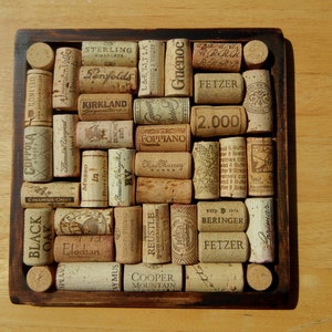 May include: A square wooden frame filled with a mosaic of used wine corks. The corks are arranged in a grid pattern and feature various brand names and logos, including Fetzer, Sterling Vineyards, and Guenoc.