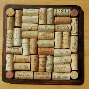 May include: A square wooden coaster with a brown frame and a cork surface. The cork surface is covered with various wine corks, each with a different brand name printed on it. The cork names include: Cambria, Catena Zapata, Charles Shaw, Sterling Vineyards, Coppola, Ruffino, Red Newt, Justin, Trellis Vineyards, Alamos, 14 Hands, William Hill, Cline, Campo Viejo, Morgan, and Seghersio.