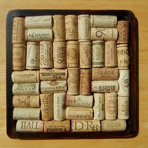 May include: A square cork trivet with a wooden frame. The trivet is filled with wine corks, each with a different winery label. Some of the labels include: King Estate, 14 Hands, Sobon, Foxbrook, Columbia Crest, Castle Rock, ELOHR, Rioja, Yorkville, Charles Shaw, Trac, Sterling, Two Vines, Bogle, Edna Valley Vineyard, 91 Cellars, Chaigne, Byron, Show, Chalone, Memoro, Francis Ford Coppola, Hall, D.R., Mis En.
