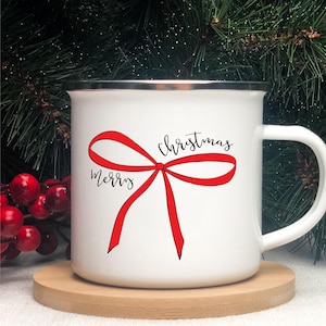 Könnte beinhalten: Weißer Emaillebecher mit rotem Schleifendesign und den Worten "Merry Christmas" in schwarzer Schrift. Der Becher hat einen silbernen Rand und einen weißen Griff. Der Becher steht auf einem Holzuntersetzer.