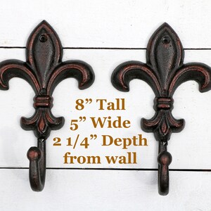 May include: Two black cast iron fleur-de-lis hooks with a brown patina finish. Each hook is 8 inches tall, 5 inches wide, and projects 2 1/4 inches from the wall.