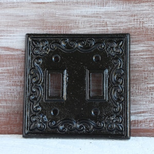 May include: Black, ornate double light switch cover with a floral and scroll design. The square cover has two rectangular openings for the switches and two screw holes. The decorative border adds a vintage or antique aesthetic.