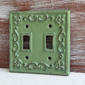 May include: A green metal light switch cover with an ornate design. The cover has two switches and is designed to fit a standard light switch.